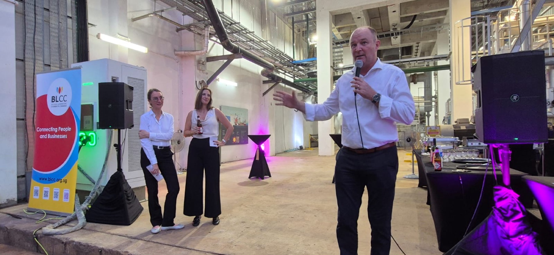 A speaker addressing the room at Powering the AI Revolution, with two colleagues listening beside a BLCC banner